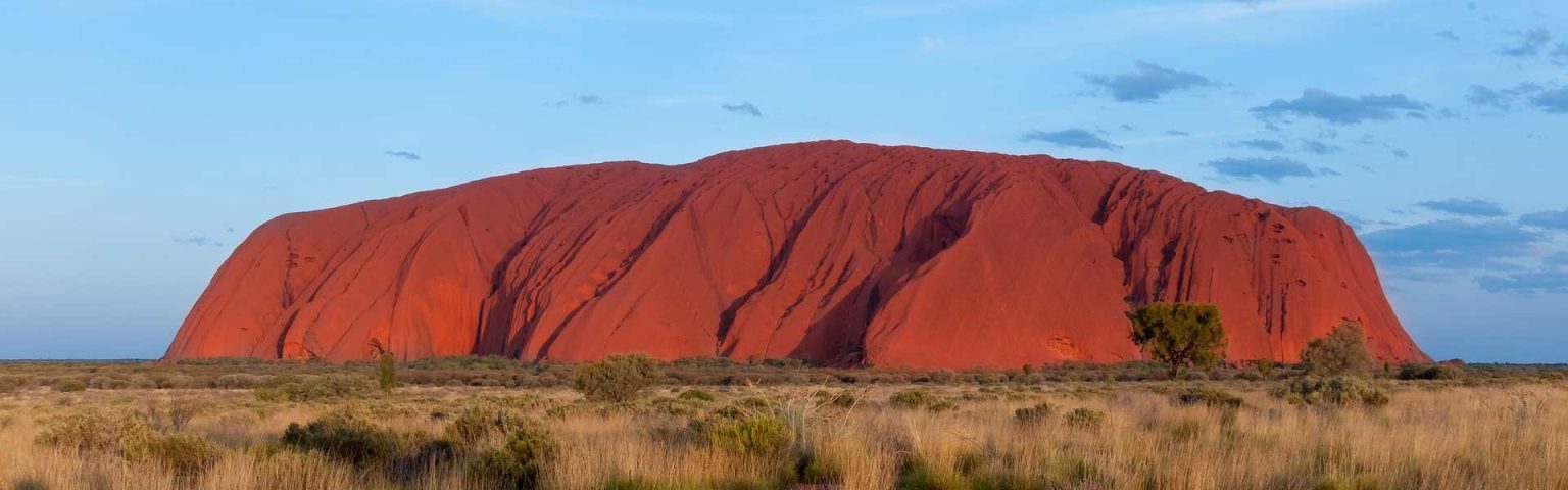 Uluru, una enorme formación rocosa de arenisca roja, se eleva sobre una pradera llana bajo un cielo azul con nubes dispersas en el interior de Australia. Se ven escasos arbustos y árboles, una escena icónica para quienes sueñan con estudiar o trabajar en Australia.